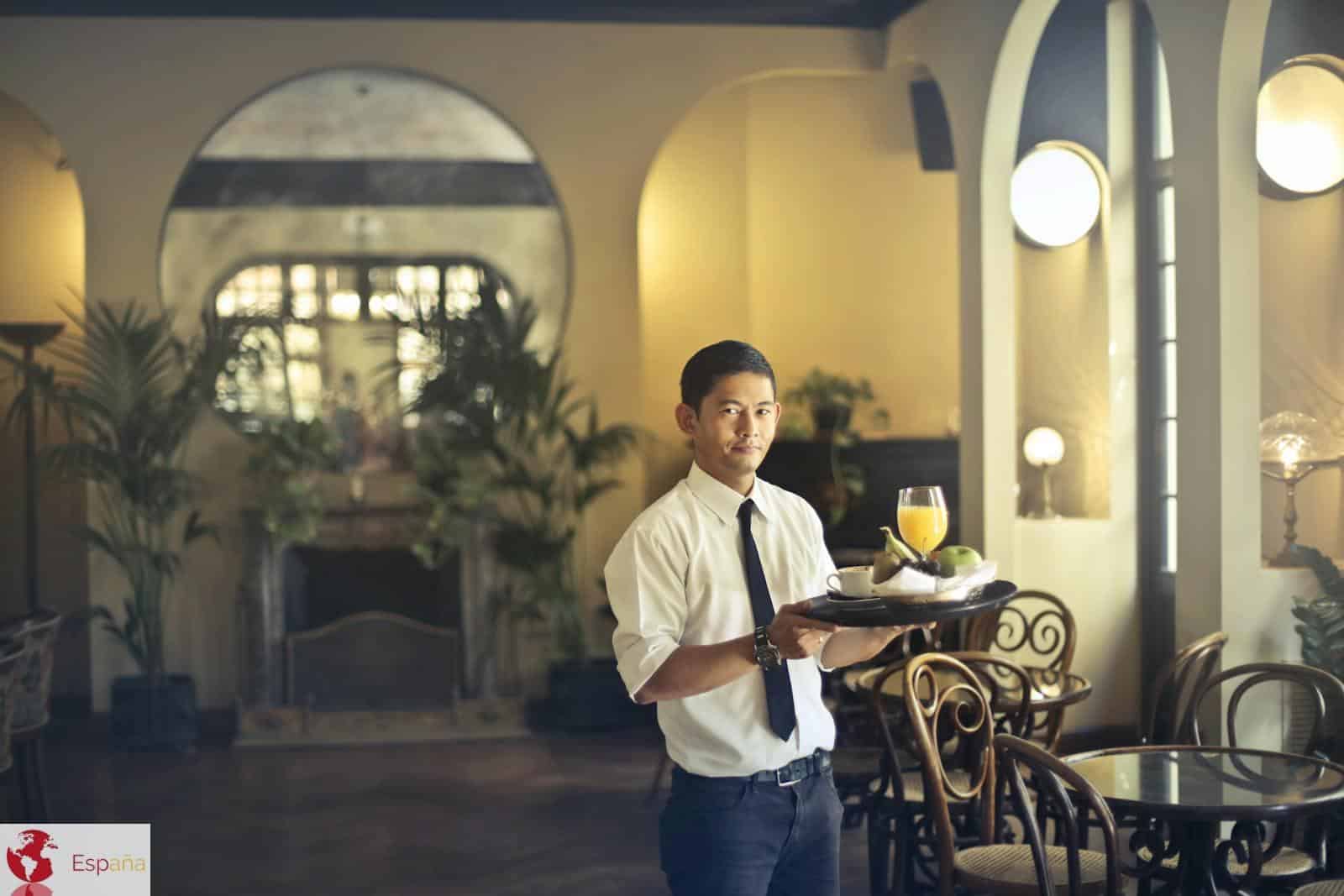 Confident young ethnic waiter in elegant clothes holding tray with food and drinks and looking at camera while serving tables in stylish restaurant